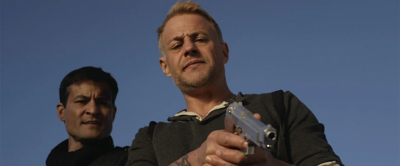 The Condemned 2 (2015) movie still: medium — Two men stand outdoors against a clear blue sky during the day. The primary subject holds a silver…
