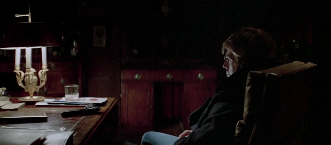 Three Days of the Condor (1975) movie still: medium — A dark study features a wooden desk holding a lamp, papers, and a pistol. A man sits in a…