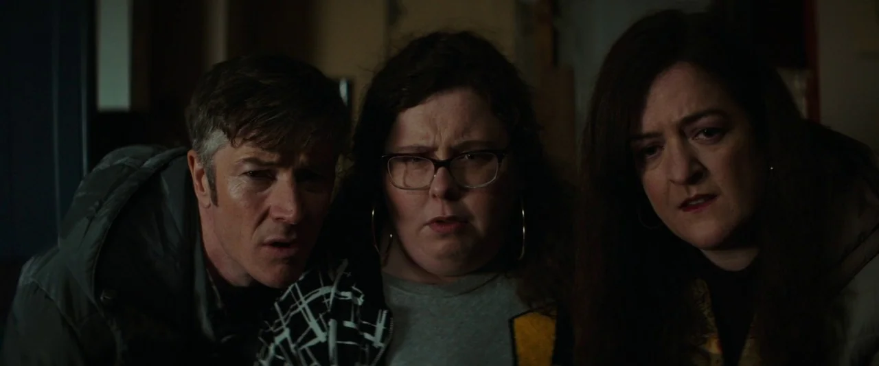 Extra Ordinary (2019) movie still: medium — Three people stand closely together inside a darkened room with soft lighting. They gaze forward with…