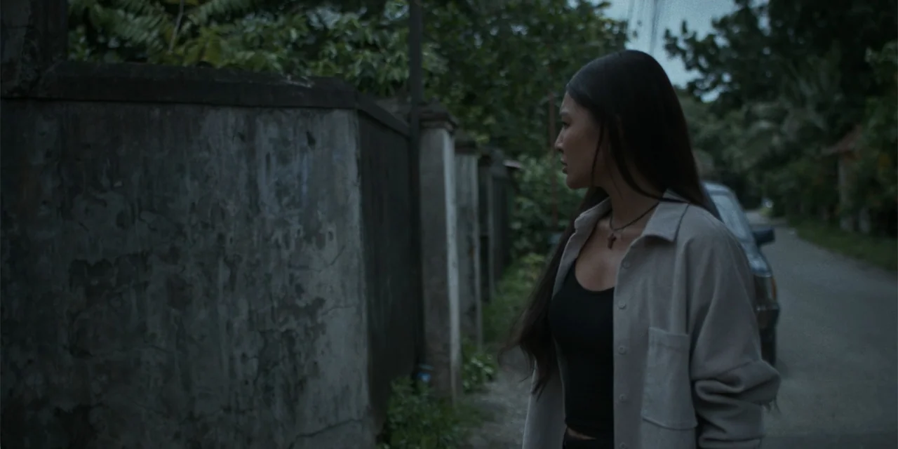 Nokturno (2024) movie still: medium — An outdoor narrow paved path lined by a tall weathered concrete boundary wall. A woman in a light grey shirt…