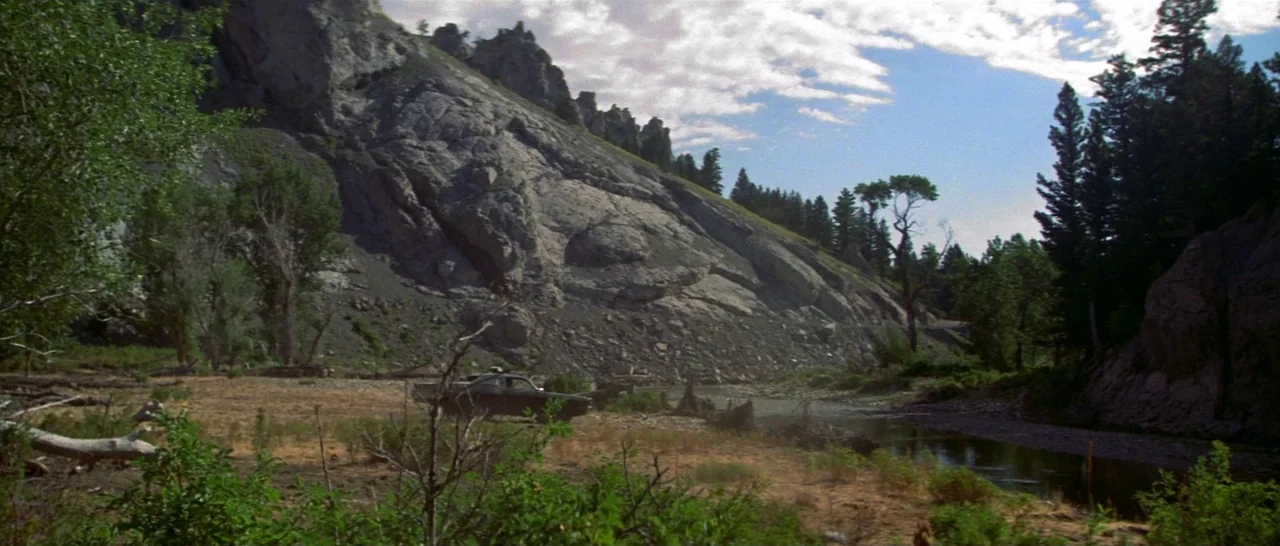 Thunderbolt & Lightfoot (1974) movie still: wide — A dark sedan sits on a rocky riverbank beneath a large steep mountain slope. Tall pine trees line…