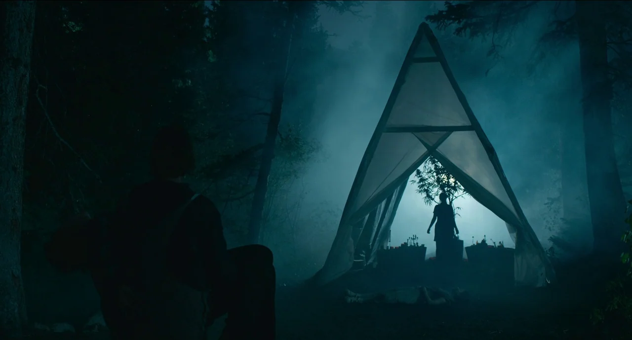Trim Season (2023) movie still: wide — A dark, misty woodland clearing contains a glowing triangular tent and dense trees. A person sits in the…