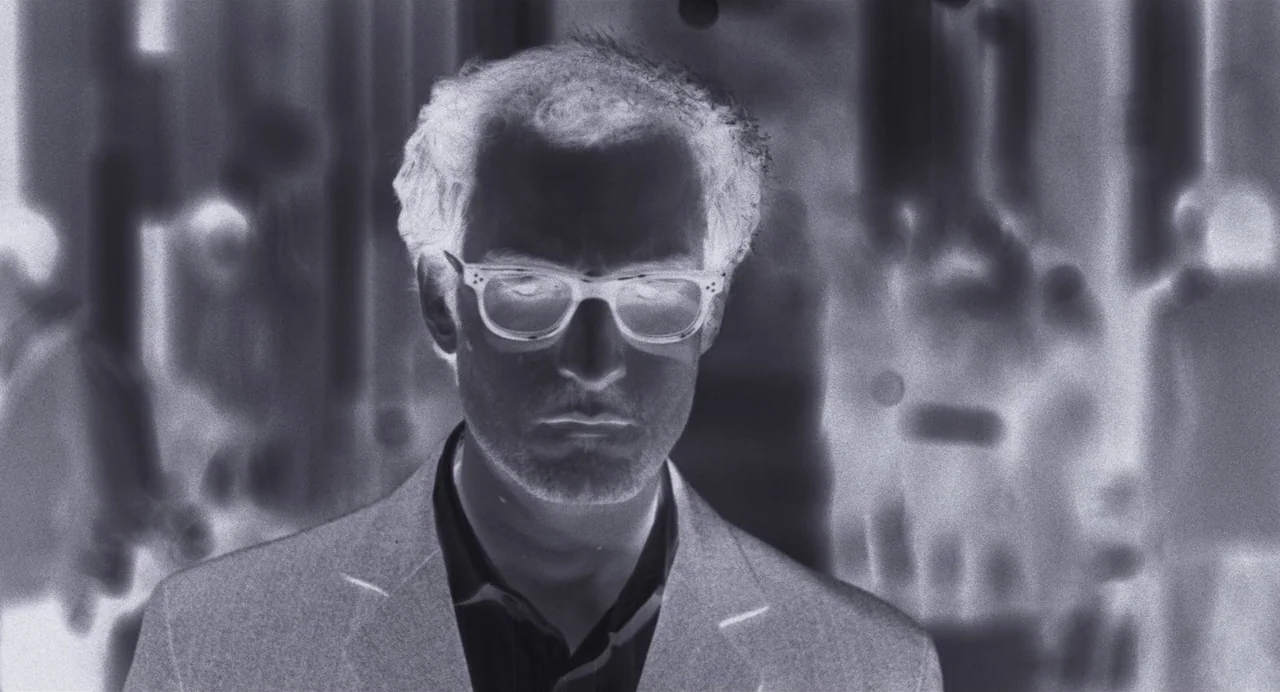 Godard Mon Amour (2015) movie still: closeup — A man walks through a crowded urban area rendered in a negative film effect. The surroundings appear…