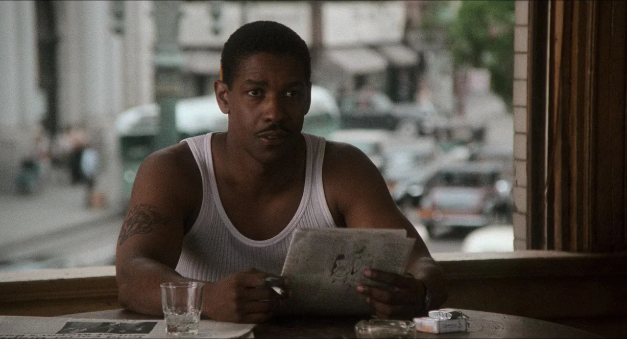 Devil In A Blue Dress (1995) movie still: medium — A man sits in a booth inside a dimly lit room overlooking a city street. He holds a newspaper in…