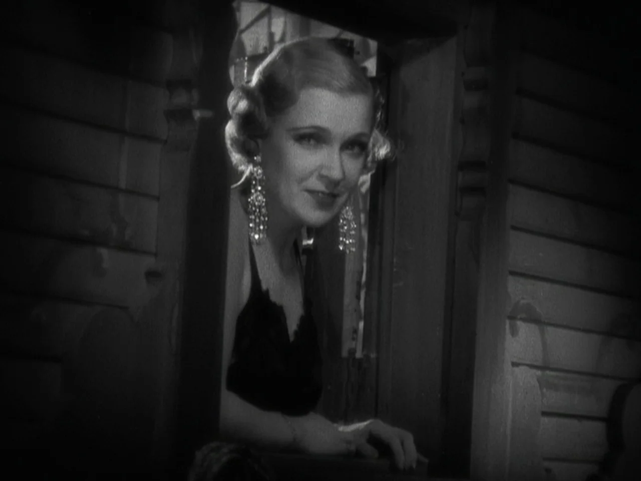 Freaks (1932) movie still: medium — A woman leans through a dark wooden window frame against a plain wall. She wears long dangling crystal earrings…