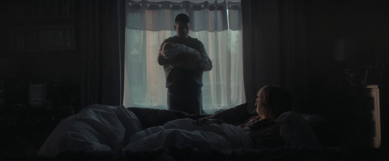 Ali & Ava (2021) movie still: medium — A dimly lit room with a bed and a window covered by curtains. A man holding a baby looks down at a woman…