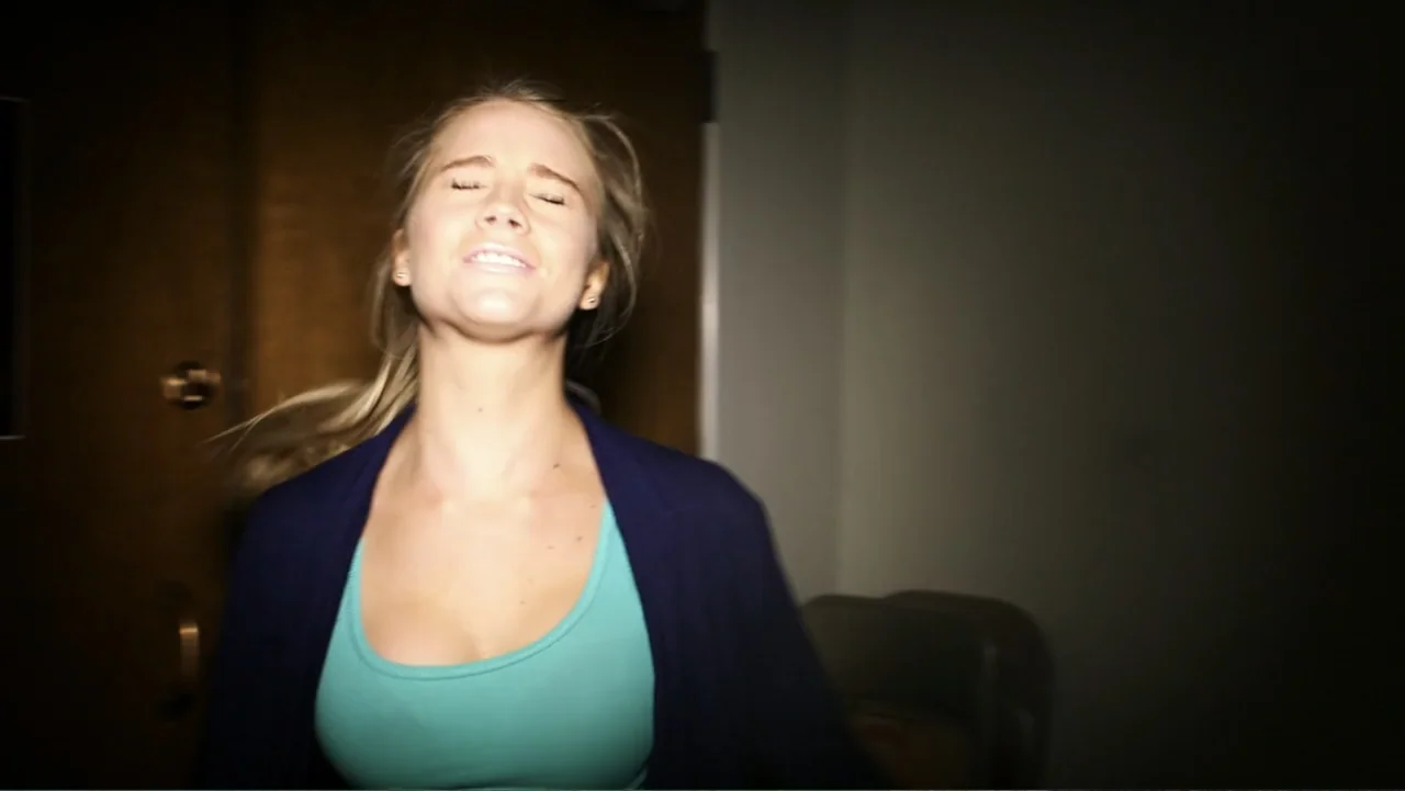 The Gallows (2015) movie still: medium — A dimly lit indoor corridor features a dark wooden door and neutral walls. A young blonde woman moves…