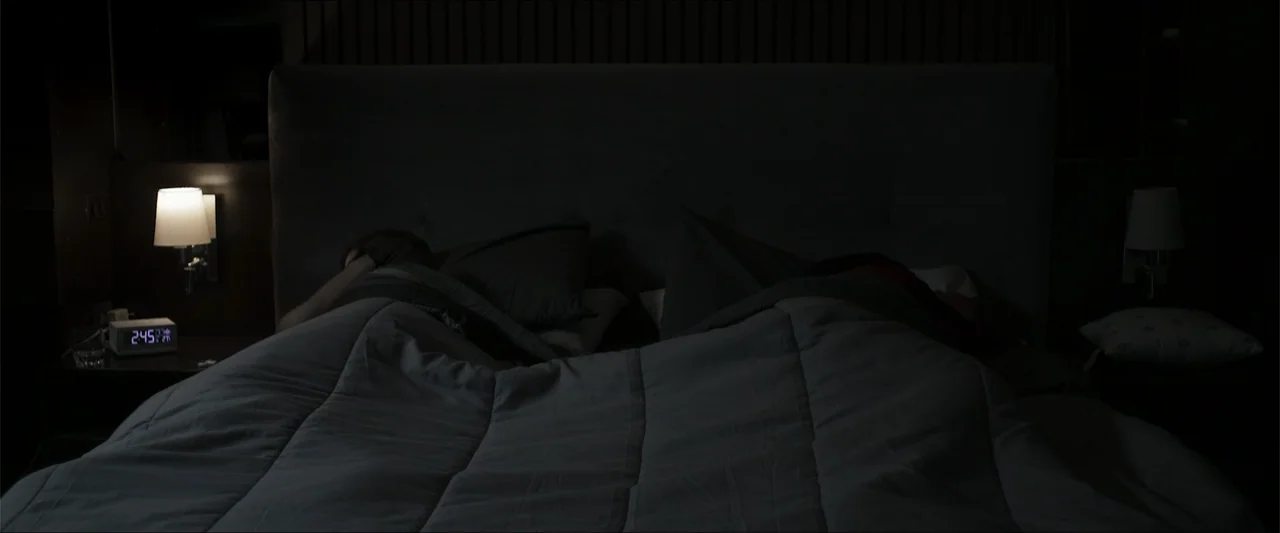 Los habitantes (2023) movie still: medium — A dimly lit bedroom features a large bed with grey linens and pillows. Two small bedside lamps and a…