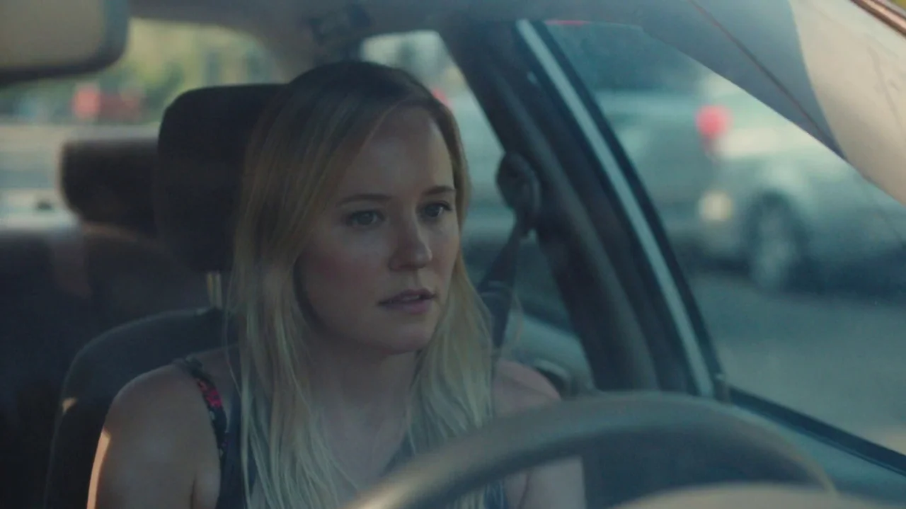 Saint Frances (2019) movie still: medium — A woman drives a car during the day with blurred traffic visible through windows. The driver holds the…