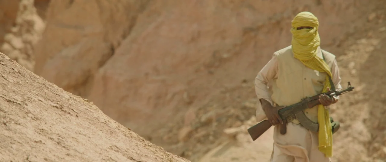 Timbuktu (2014) movie still: medium — A dry, sun-scorched canyon floor with sheer rock faces rising in the background. A man stands in the…
