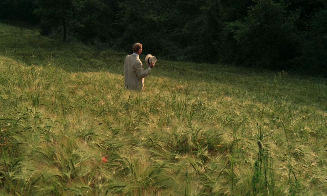 A Room With A View (1985) movie still: wide — A field of tall ripening grass bordered by dense trees in the background. A man in a beige linen suit…