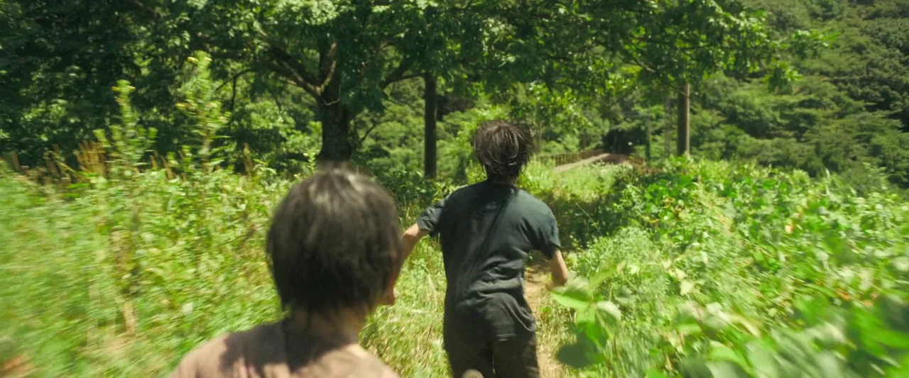 Monster (2023) movie still: medium — A narrow dirt path cuts through thick green vegetation and tall trees under sunlight. Two youths run along the…