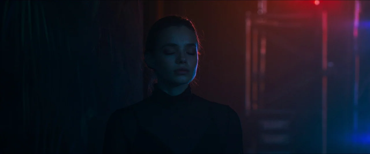Birds of Paradise (2021) movie still: medium — A dark space features a woman standing in front of a blurred out-of-focus background. She stands…