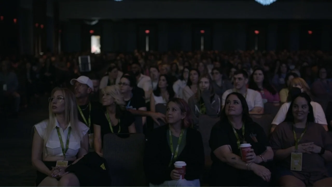 Predators (2010) movie still: medium — A darkened interior conference hall features rows of seated individuals facing forward. The attendees wear…