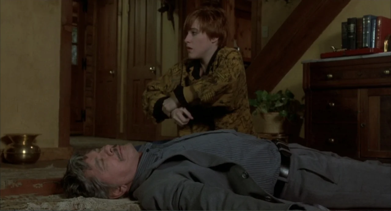 Murphy's Law (2009) movie still: medium — A dimly lit room features a hardwood floor and wooden furniture in the background. A woman kneels beside a…
