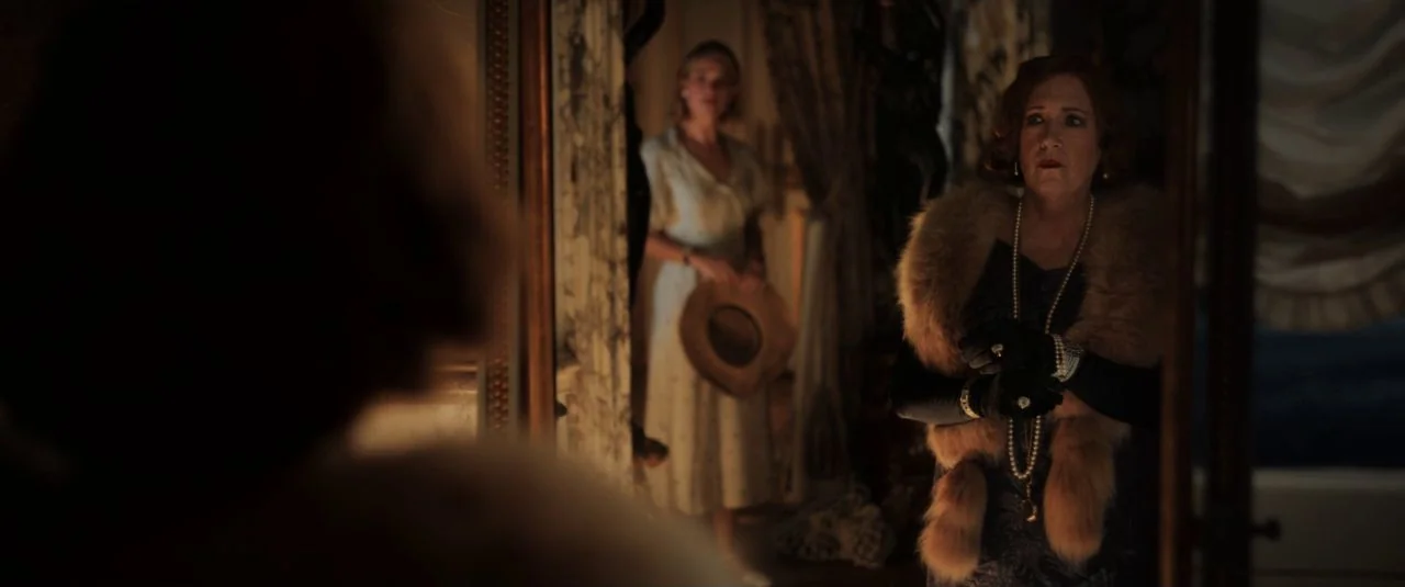 Rebecca movie still: medium — A dimly lit room with ornate drapery and a large antique mirror reflecting two women. An older woman in a fur stole…