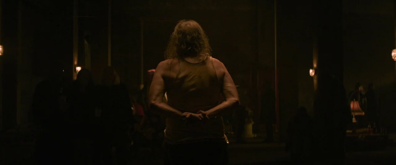 Pig (2021) movie still: medium — A dark, sparsely lit room with indistinct figures standing in the deep shadows. A man stands in the foreground,…