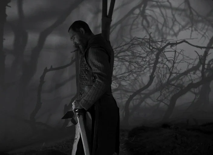 The Tragedy of Macbeth (2021) movie still: medium — A dense, fog-filled forest with bare, twisted tree limbs in the background. A bearded man stands…