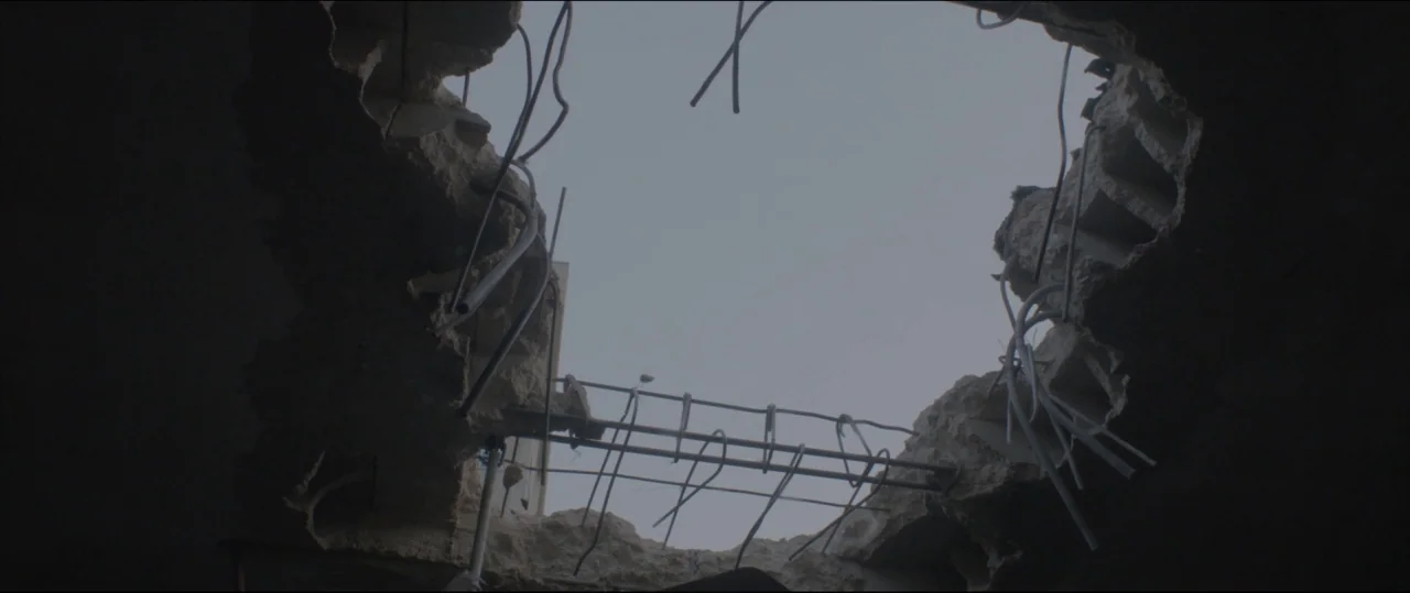 Under The Shadow (2016) movie still: establishing — A jagged hole in a concrete ceiling reveals an overcast sky above. Exposed metal rebar protrudes…
