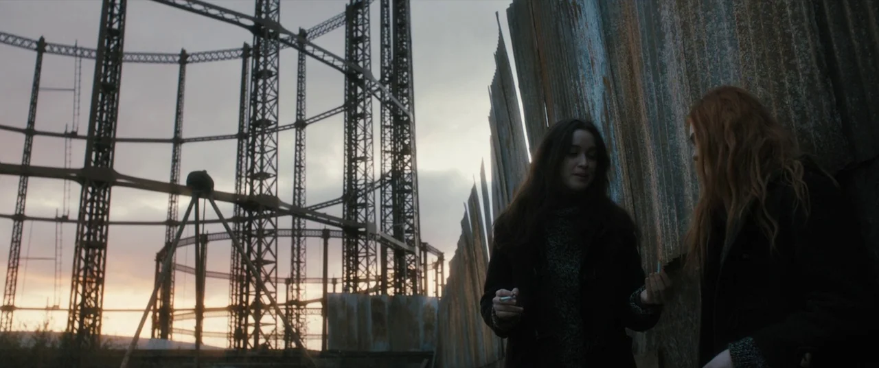 Ginger & Rosa (2012) movie still: medium — A desolate industrial area features a large iron gasometer frame under a cloudy sky. Two young women…