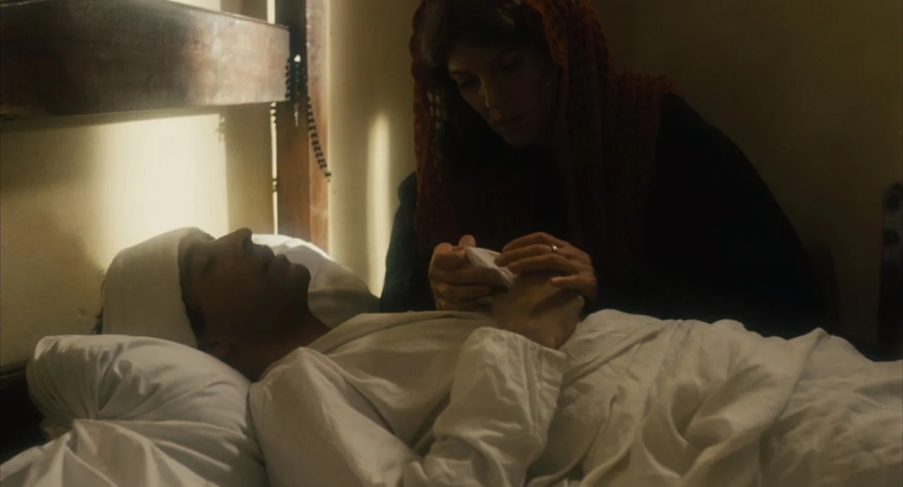 Camila (1984) movie still: medium — A spare bedroom features plain light-colored walls and a wooden bed frame. A woman sits beside a man in the bed,…