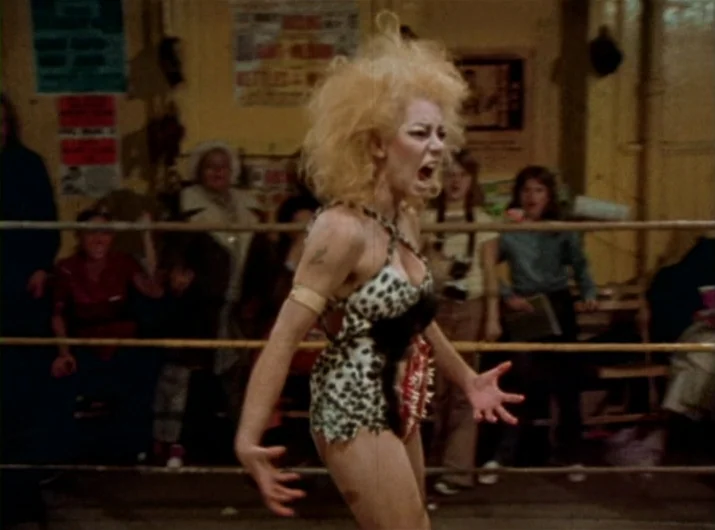 Desperate Living (1977) movie still: medium — A dimly lit indoor space with ropes forming a makeshift wrestling ring enclosure. A blonde woman with…
