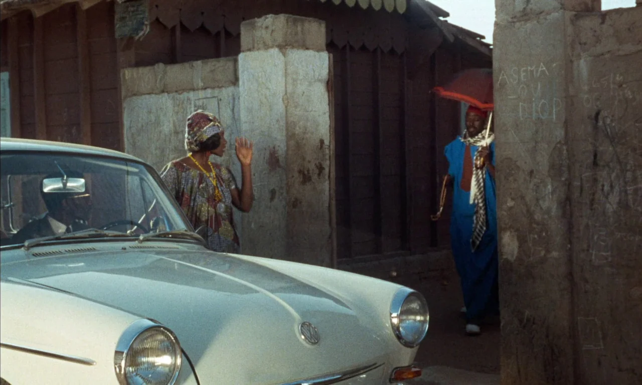 Mandabi (1968) movie still: medium — A street corner features a parked vintage Volkswagen sedan near a concrete wall structure. A woman stands…