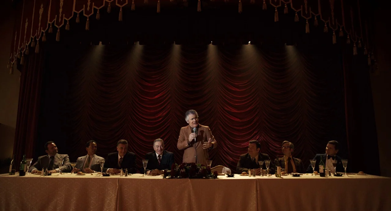 The Irishman (2019) movie still: wide — A banquet hall features a long table set for a formal dinner before red curtains. A group of men sit at the…
