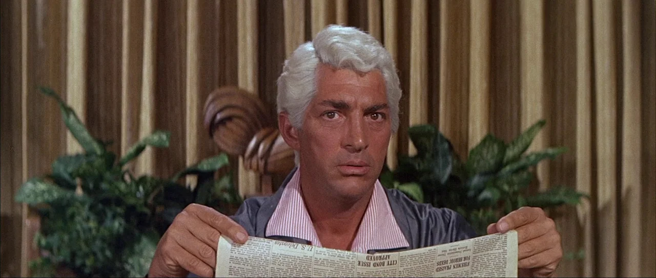What A Way To Go! (1964) movie still: medium — A man sits before a wall of hanging fabric curtains and two green houseplants. He holds a…