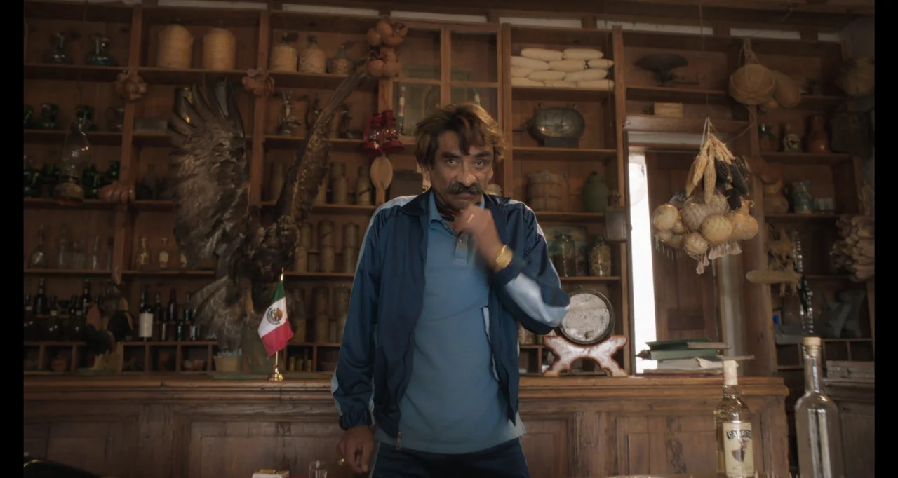 National Family (2023) movie still: medium — A cluttered vintage shop interior features wooden shelves filled with various jars and artifacts. A man…