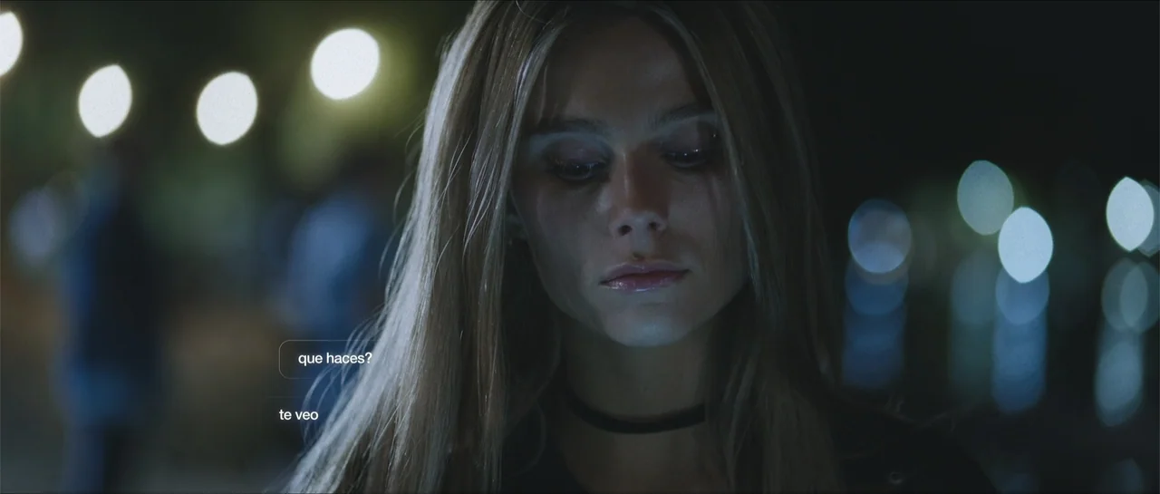 Nahir (2024) movie still: closeup — A woman stands outdoors at night with blurry light bokeh in the background. She looks downward while text…