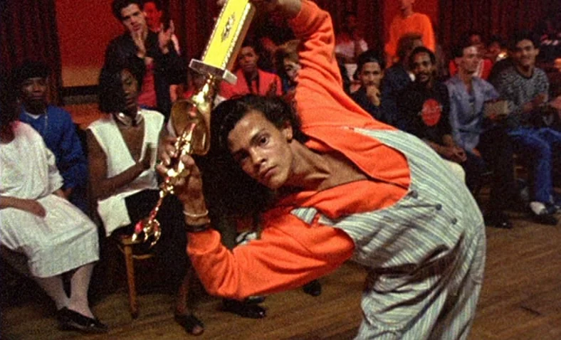 Paris Is Burning (1990) movie still: medium — A dark ballroom with rows of people seated in the background on chairs. A young man in an orange…