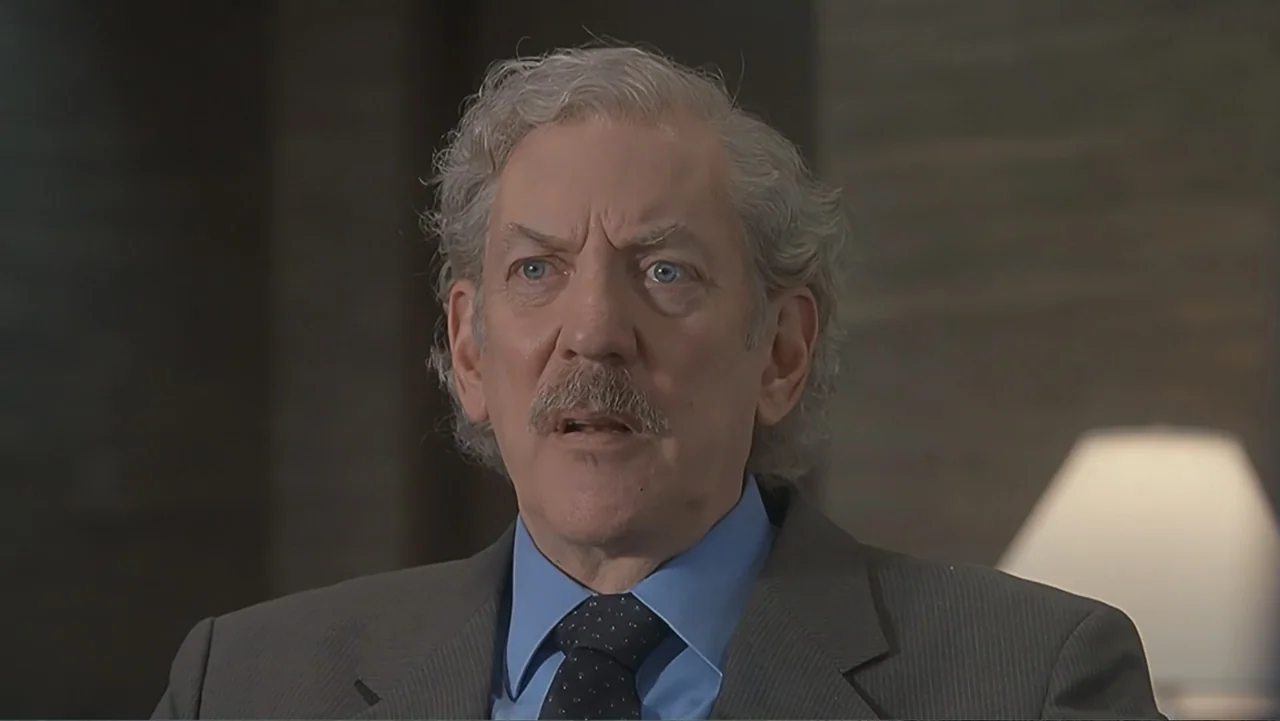 The Art of War (2000) movie still: closeup — A professional interior space with blurred wall textures and a lamp in the distance. An older man in a…