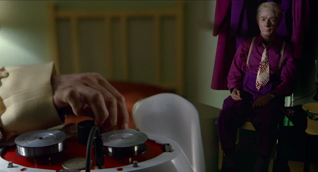 Dick Tracy (1990) movie still: medium — A dimly lit room contains a person seated near a clothing rack. In the foreground a hand manipulates a…