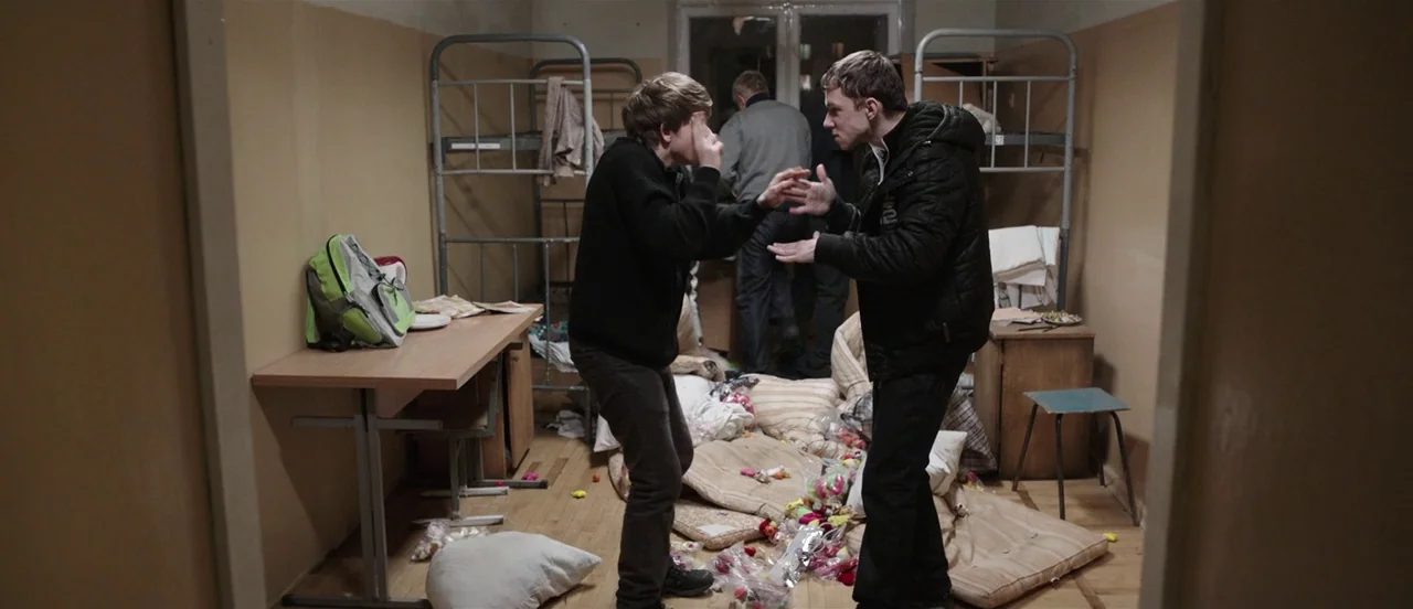 The Tribe (2014) movie still: fullbody — A cramped and cluttered dormitory room with discarded mattresses and trash scattered on the floor. Two…