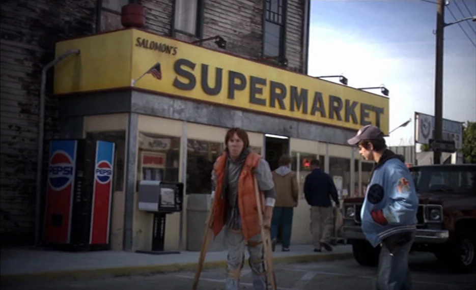 Dear Wendy (2005) movie still: fullbody — The exterior of Salomon's Supermarket features a yellow sign and weathered wood paneling. Two teenagers…