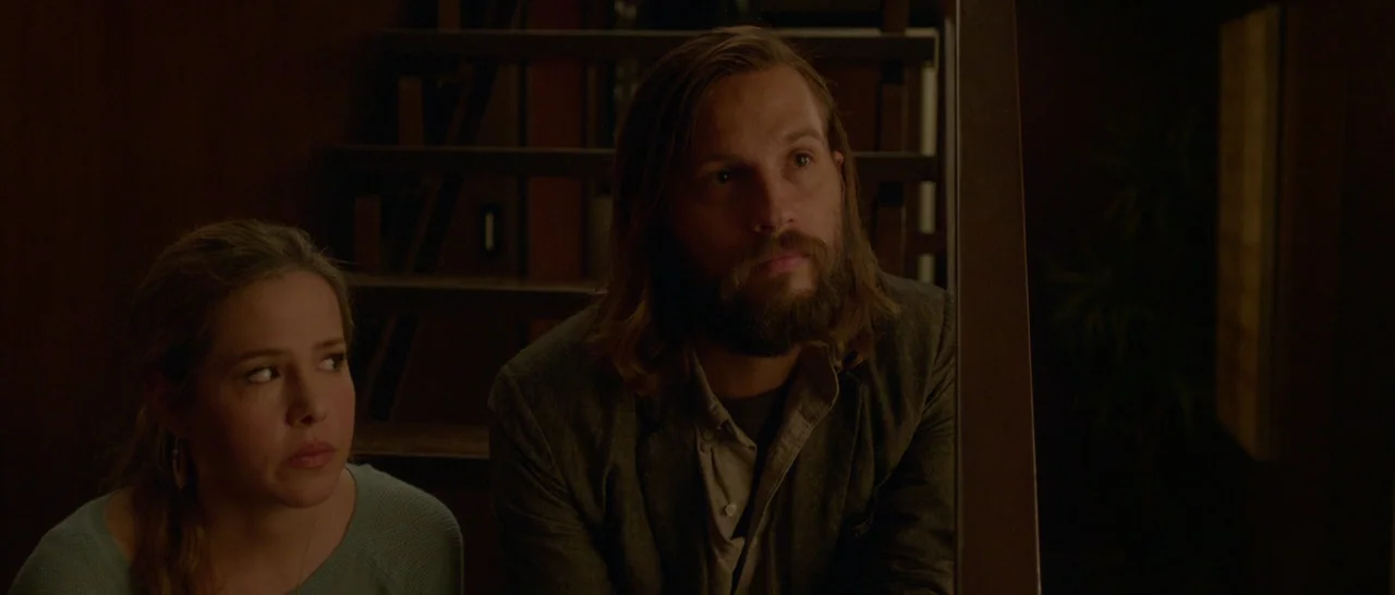 The Invitation (2015) movie still: medium — A dimly lit residential stairwell with dark wooden steps and walls at night. A bearded man and a woman…