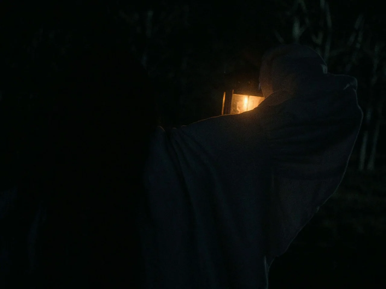 First Cow (2019) movie still: medium — A dark area with a figure partially visible in the shadows. The figure holds a single glowing lantern while…