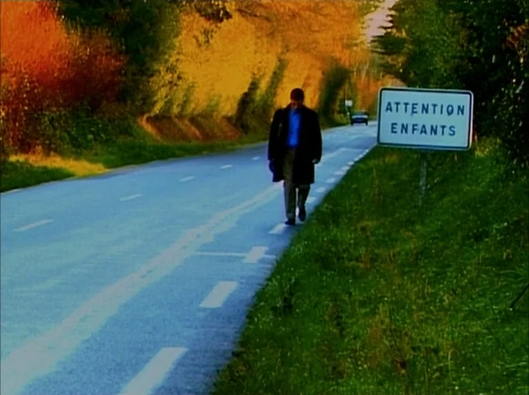 Eloge de L’amour (2001) movie still: wide — A rural paved road is lined with vibrant autumn trees and grassy embankments. A man in a dark coat walks…