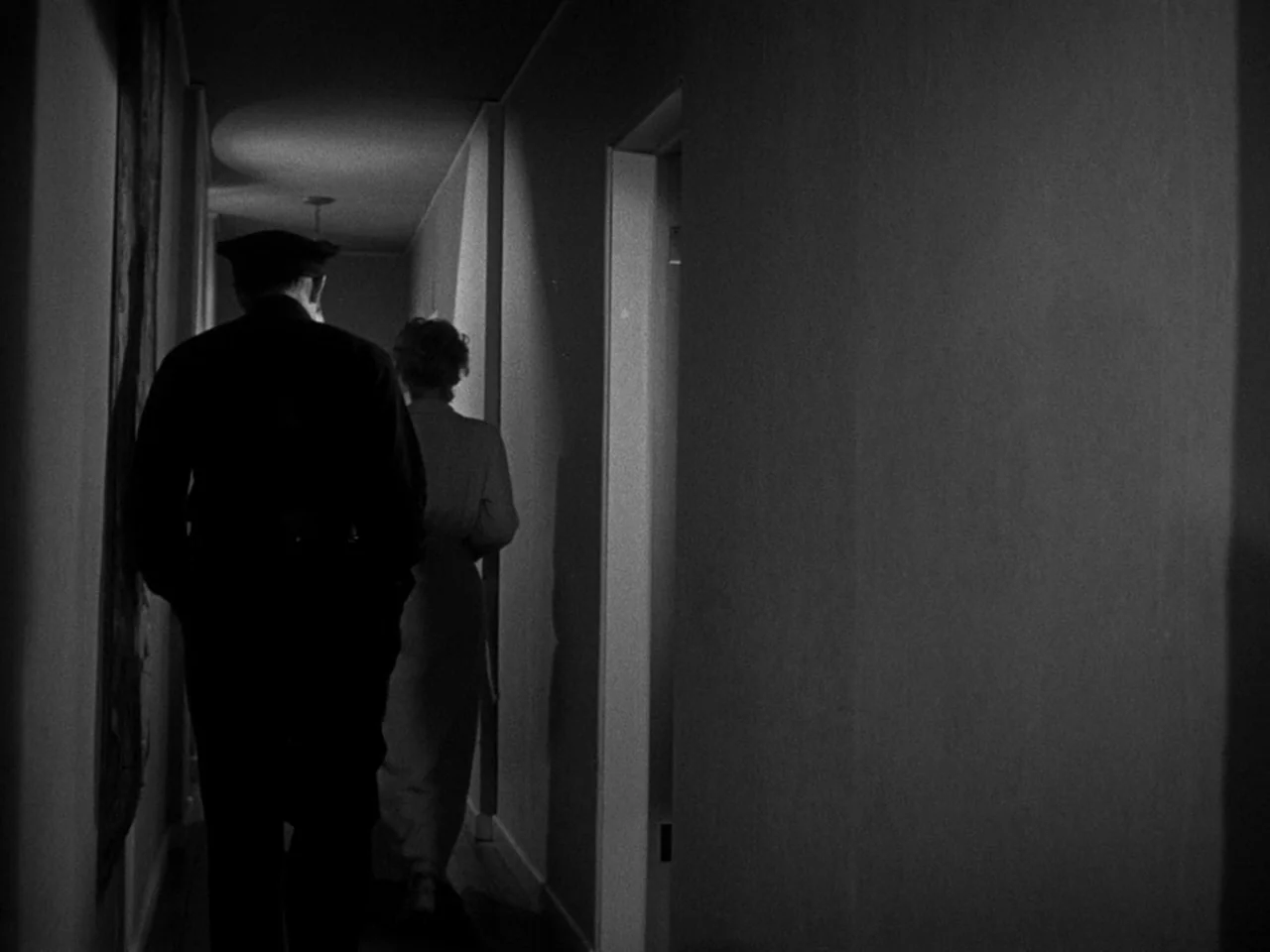 The Prowler (1951) movie still: fullbody — A dark residential hallway with plain walls and a single door frame visible. A man in a police uniform…