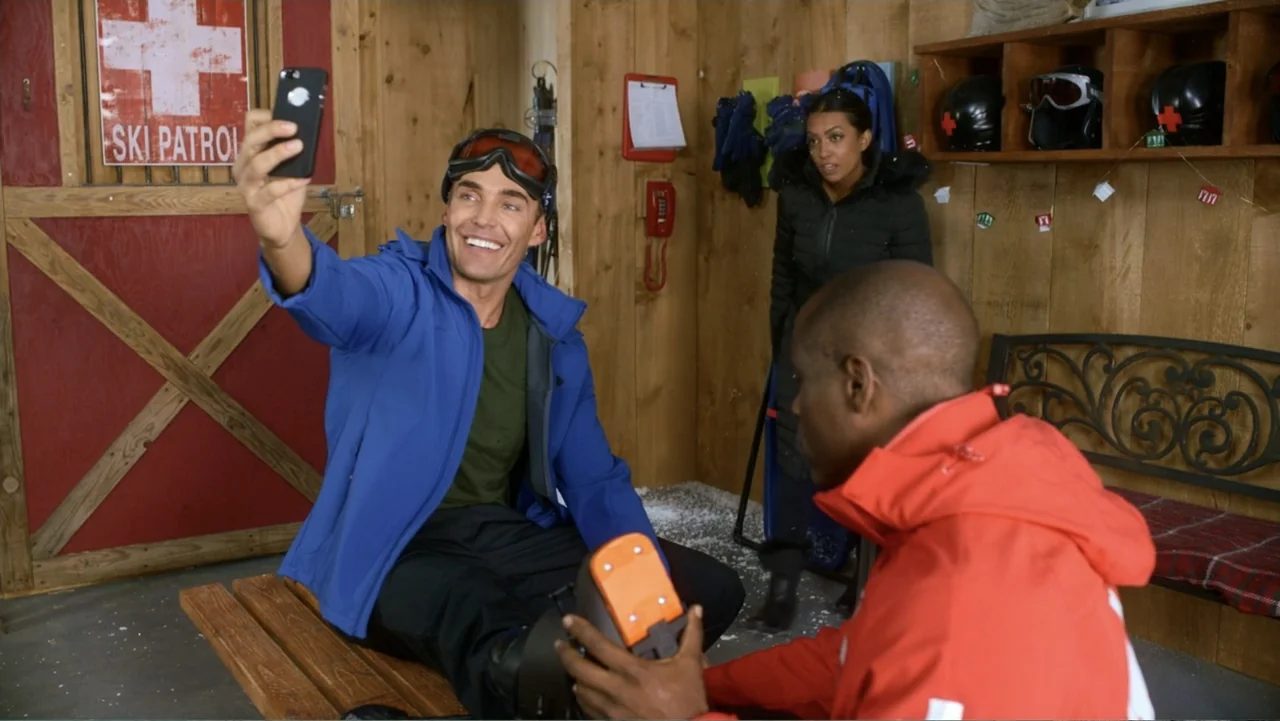 Christmas with a Prince (2018) movie still: medium — A wooden ski patrol office features paneled walls and a red wooden door. A man sits on a bench…