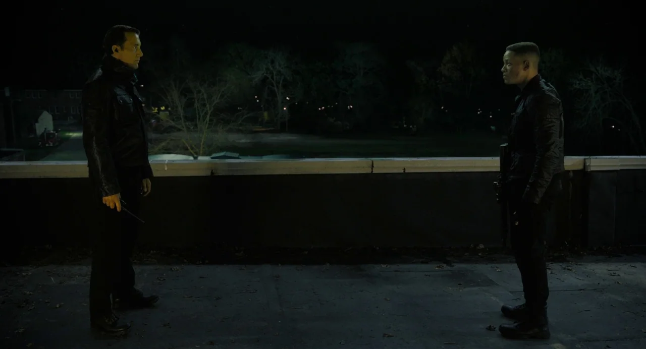 Gemini Man (2019) movie still: fullbody — A wide shot shows two men standing on a dark flat roof at night. The pair stand in profile, separated by…