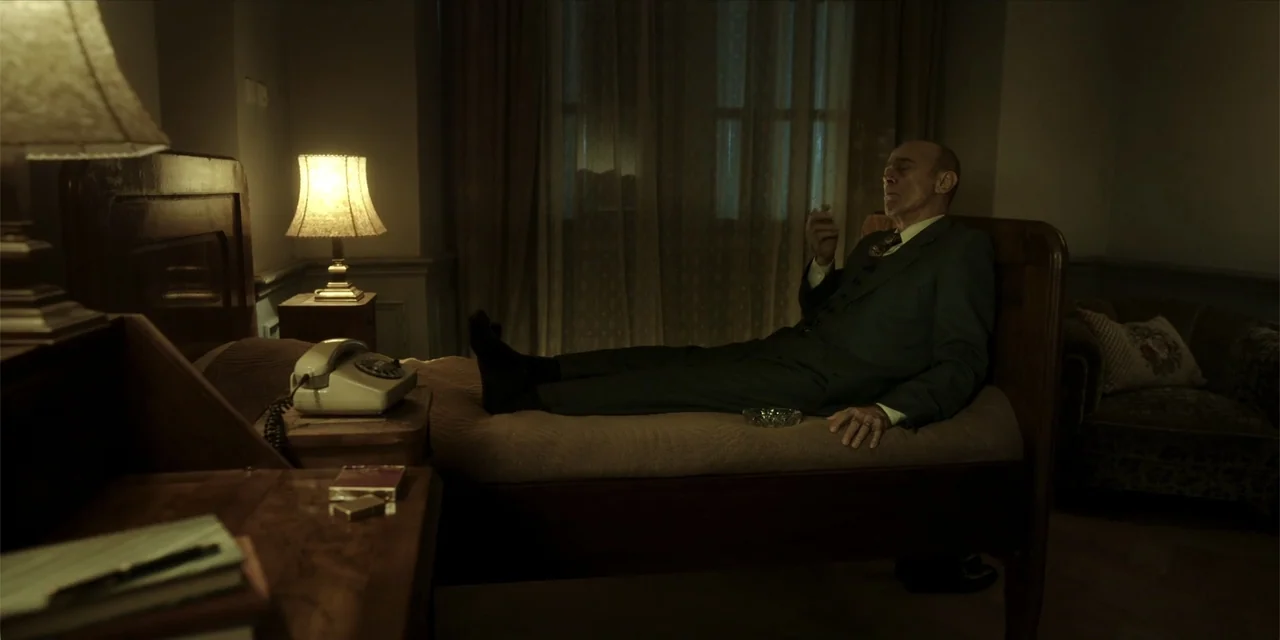 Oslo (2021) movie still: medium — A dimly lit 1960s-style hotel room features a bed, a desk, and a warm lamp. An elderly man in a suit lies on the…