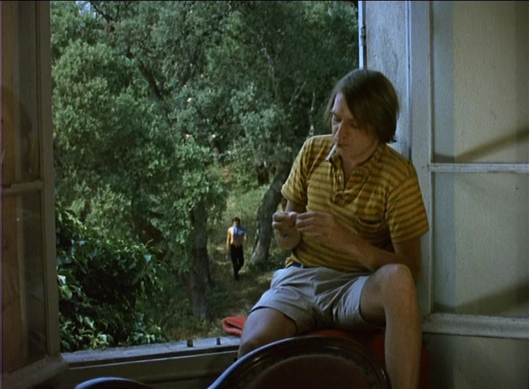 La Collectionneuse (1967) movie still: medium — A man sits on a ledge of an open window overlooking dense foliage. Another shirtless man walks…