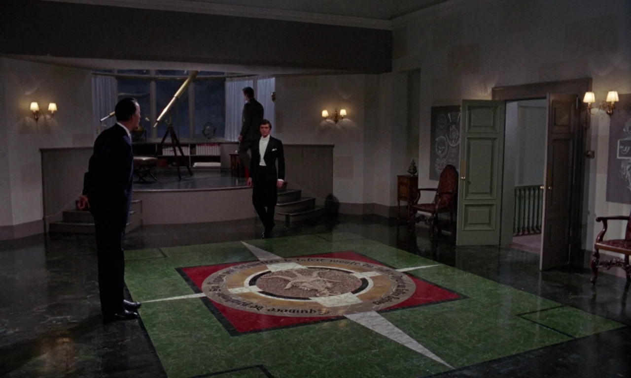 The Devil Rides Out (1968) movie still: wide — A spacious room features a large compass rose inlaid into a green marble floor. Three men are…