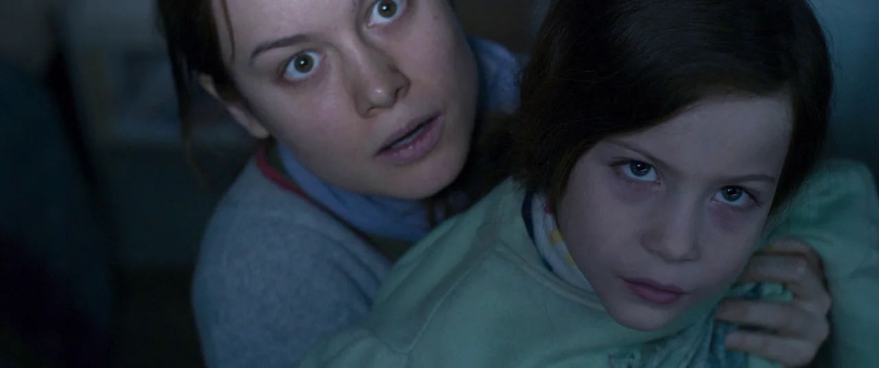 Room (2015) movie still: medium — An dimly lit interior space is occupied by an adult woman and a child. The woman embraces the child from behind…