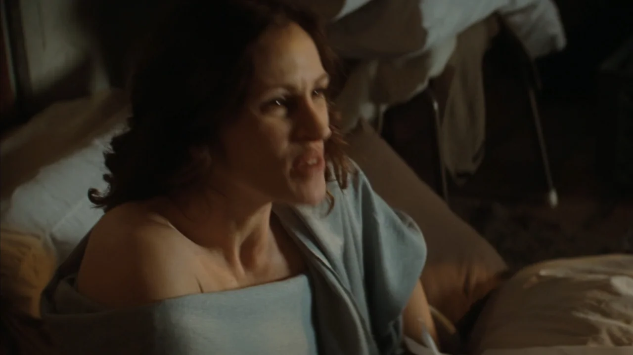 Duplicity (2009) movie still: medium — A dimly lit room contains a bed with soft white pillows and wrinkled sheets. A woman with dark wavy hair sits…