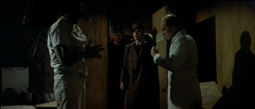 MASH (1970) movie still: fullbody — A dimly lit industrial room contains dark wooden partitions and medical supply boxes. Two men in coats and a…