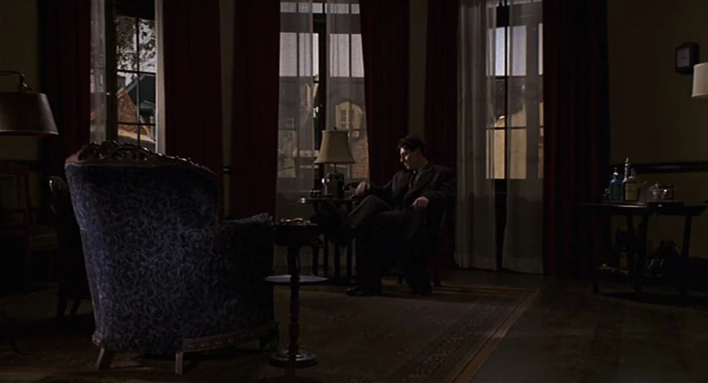 Miller’s Crossing (1990) movie still: wide — A dimly lit room features a dark patterned armchair in the foreground. A man sits in a chair in the…