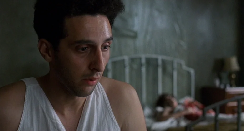 Barton Fink (1991) movie still: closeup — A dimly lit room features a metal-framed bed and dark, textured walls. A man in a white tank top sits in…